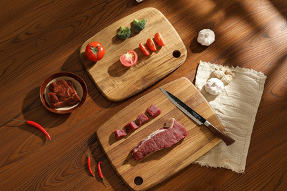 Two acacia wood cutting boards being used to prepare vegetables and meat on a kitchen table.