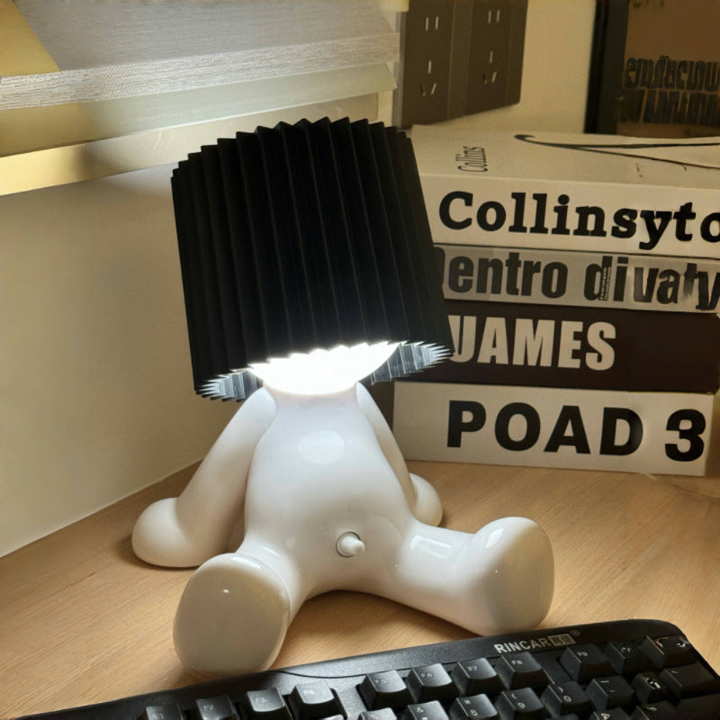 White seated character desk lamp with a glowing light and black pleated shade displayed beside books and a keyboard.