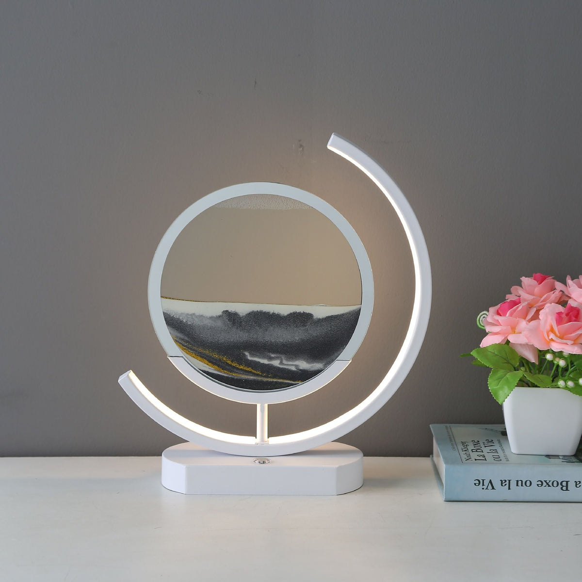 White sand art table lamp with a glowing curved frame and layered black and white sand, displayed beside pink flowers.