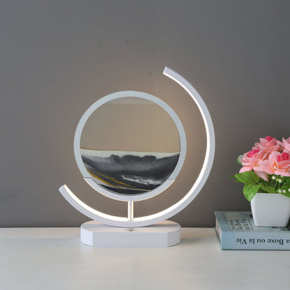 White sand art table lamp with a glowing curved frame and layered black and white sand, displayed beside pink flowers.