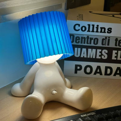 White seated character desk lamp with a glowing light and blue pleated shade displayed beside a keyboard.