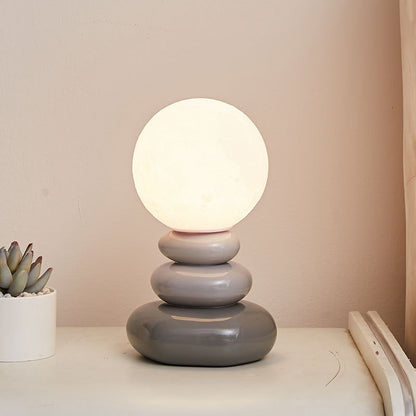 Table lamp with three stacked gray ceramic pebble shapes and an illuminated round globe shade beside a small plant.