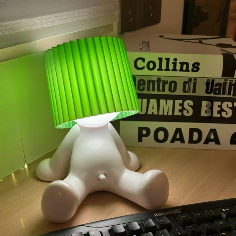 White seated character desk lamp with a glowing light and green pleated shade displayed beside books.