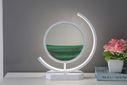 Green sand forms layered patterns inside a circular display framed by a glowing white curved table lamp.