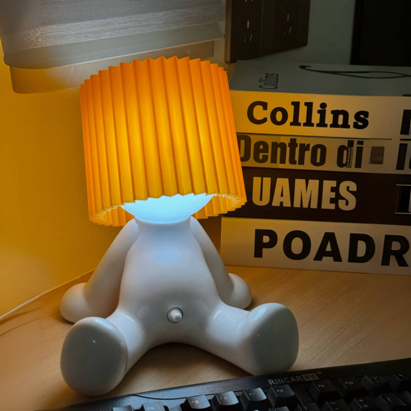 White seated character desk lamp with a glowing light and orange pleated shade on a desk.