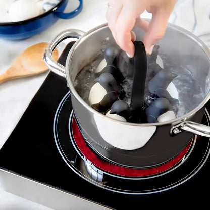 Hand holding a penguin-shaped egg holder containing eggs in a pot of bubbling water on a stovetop.