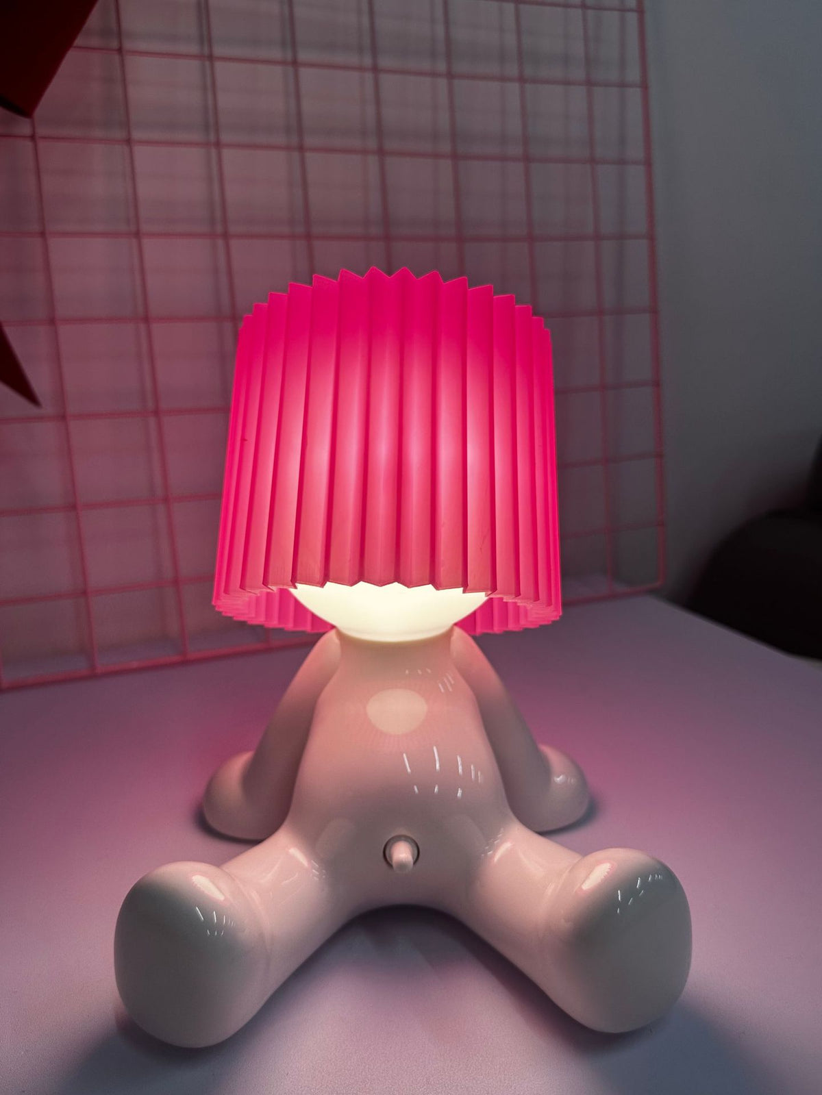 White seated character desk lamp glowing beneath a bright pink pleated shade on a tabletop.
