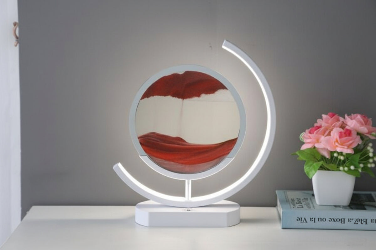 Red sand falling into layered patterns inside a round display surrounded by a glowing white curved lamp frame.