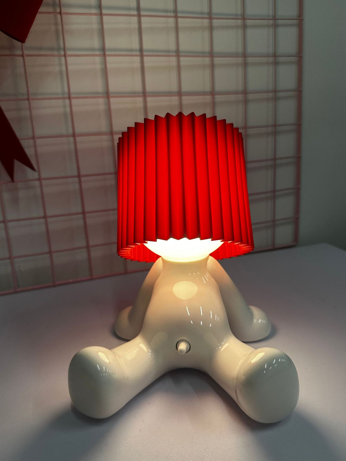 White seated character desk lamp glowing beneath a red pleated shade on a tabletop.