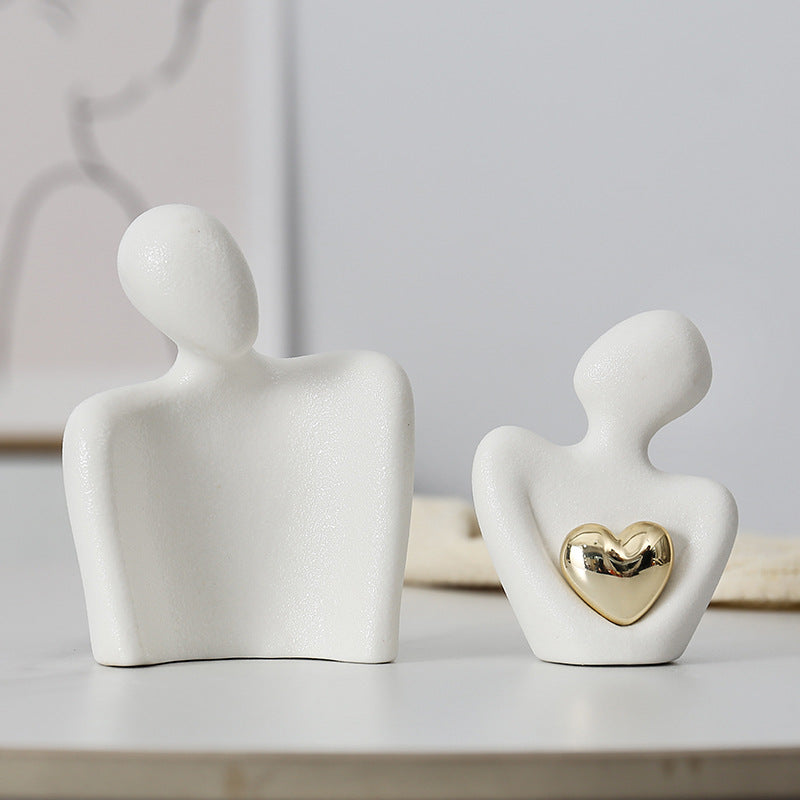 White ceramic couple sculpture shown as two separate figures, with a gold-colored heart on the smaller front figure.