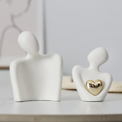 White ceramic couple sculpture shown as two separate figures, with a gold-colored heart on the smaller front figure.