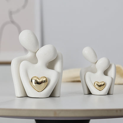 Two white ceramic couple sculptures in different sizes, each with abstract embracing figures and a gold-colored heart.