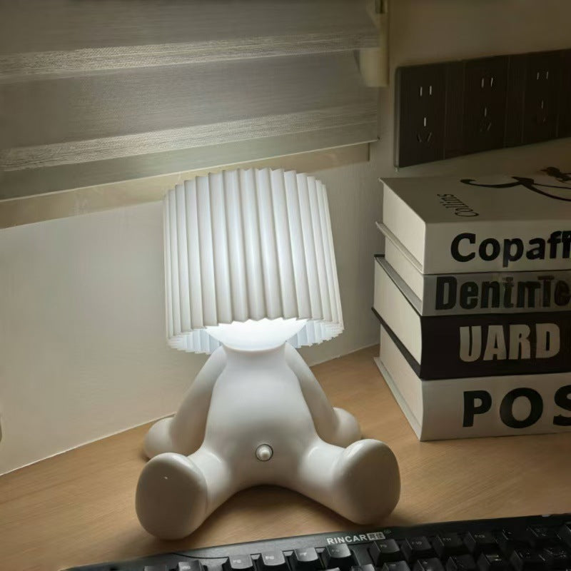 White seated character desk lamp with a glowing light and matching white pleated shade beside stacked books.