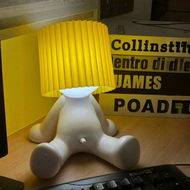 White seated character desk lamp with a glowing light and bright yellow pleated shade on a desk.