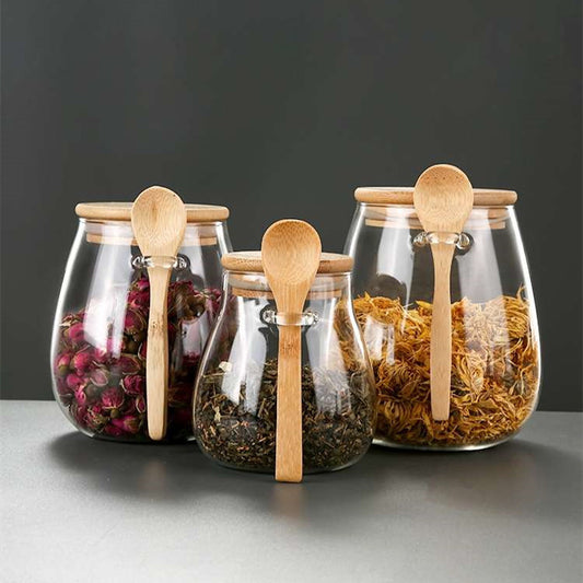 Three glass spice jars with bamboo lids and wooden spoons filled with dried herbs and spices