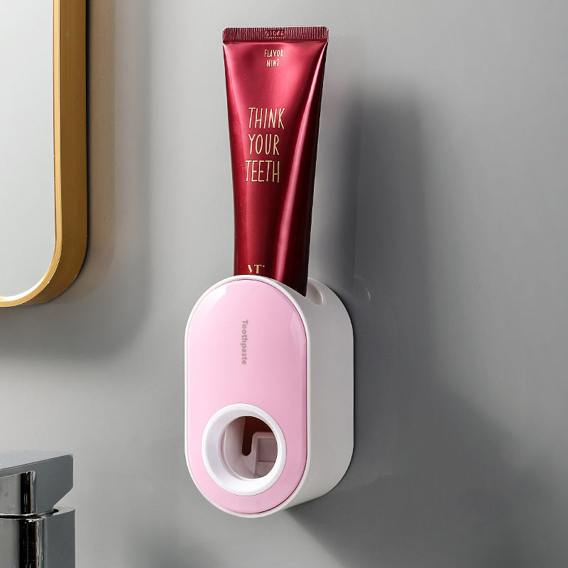 Pink and white wall-mounted toothpaste dispenser holding a toothpaste tube beside a bathroom mirror.