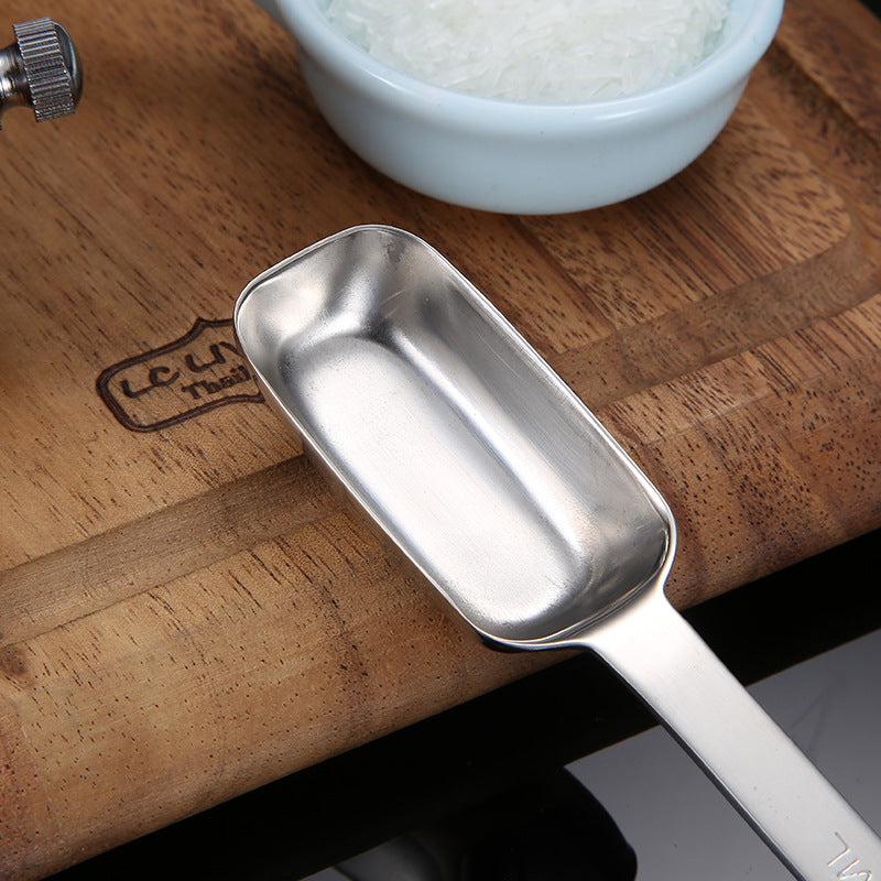 Close-up of a rectangular stainless steel measuring spoon beside a bowl of flour