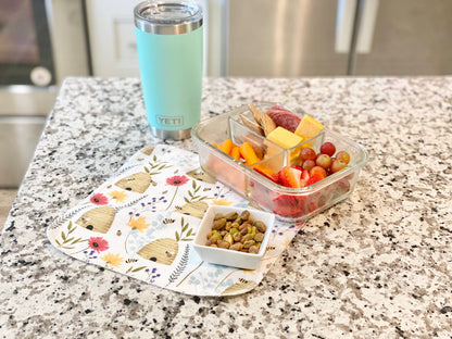 Reusable flannel paper towel with a bee and floral print used under a snack container and drink on a kitchen countertop