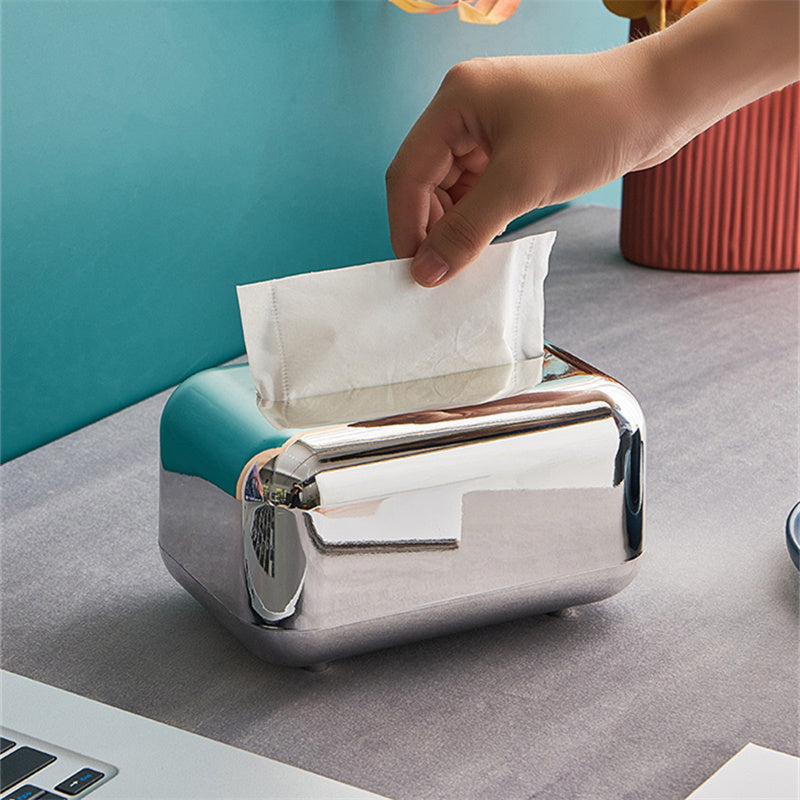 A hand pulling a tissue from a silver tissue box with rounded corners and a reflective finish.