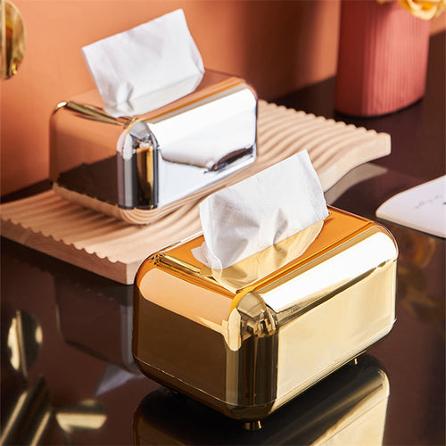 Silver and gold tissue boxes with rounded corners and reflective finishes, displayed together on a table.
