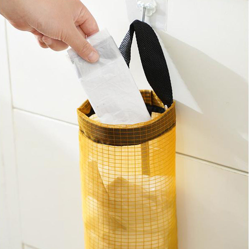 A reusable plastic bag being placed into a yellow hanging mesh bag holder mounted on a kitchen wall.